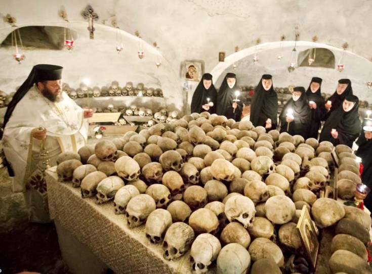 A Orthodox priest perform his religious duties in front of a table with skulls. 